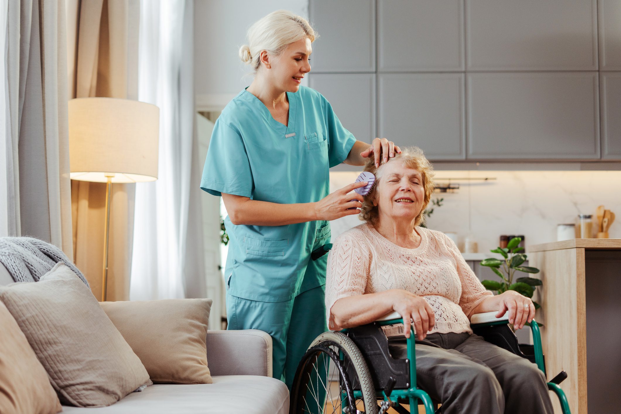 Nurse caring for a senior woman in a wheelchair at home, brushing her hair with compassion and warmth. The image shows the bond between caregiver and patient